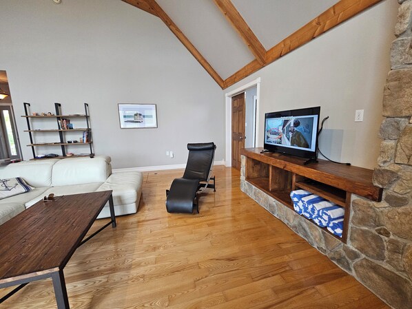 Una sala de estar con piso de madera, un sofá blanco, un sillón de cuero negro, un televisor sobre un mueble de madera y una pared de piedra.