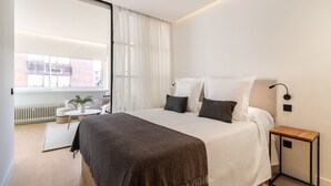 Un dormitorio moderno con una cama grande, una mesita de noche, una pequeña mesa redonda con una planta y vistas a la ciudad a través de un ventanal amplio.