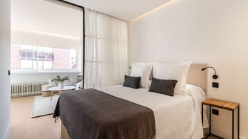 Een moderne slaapkamer met een groot bed, een nachtkastje, een klein rond tafeltje met een plant en een uitzicht op de stad door een groot raam.