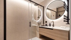 Un baño moderno con ducha envuelta en vidrio, espejo redondo, tocador de madera y lavamanos blanco.