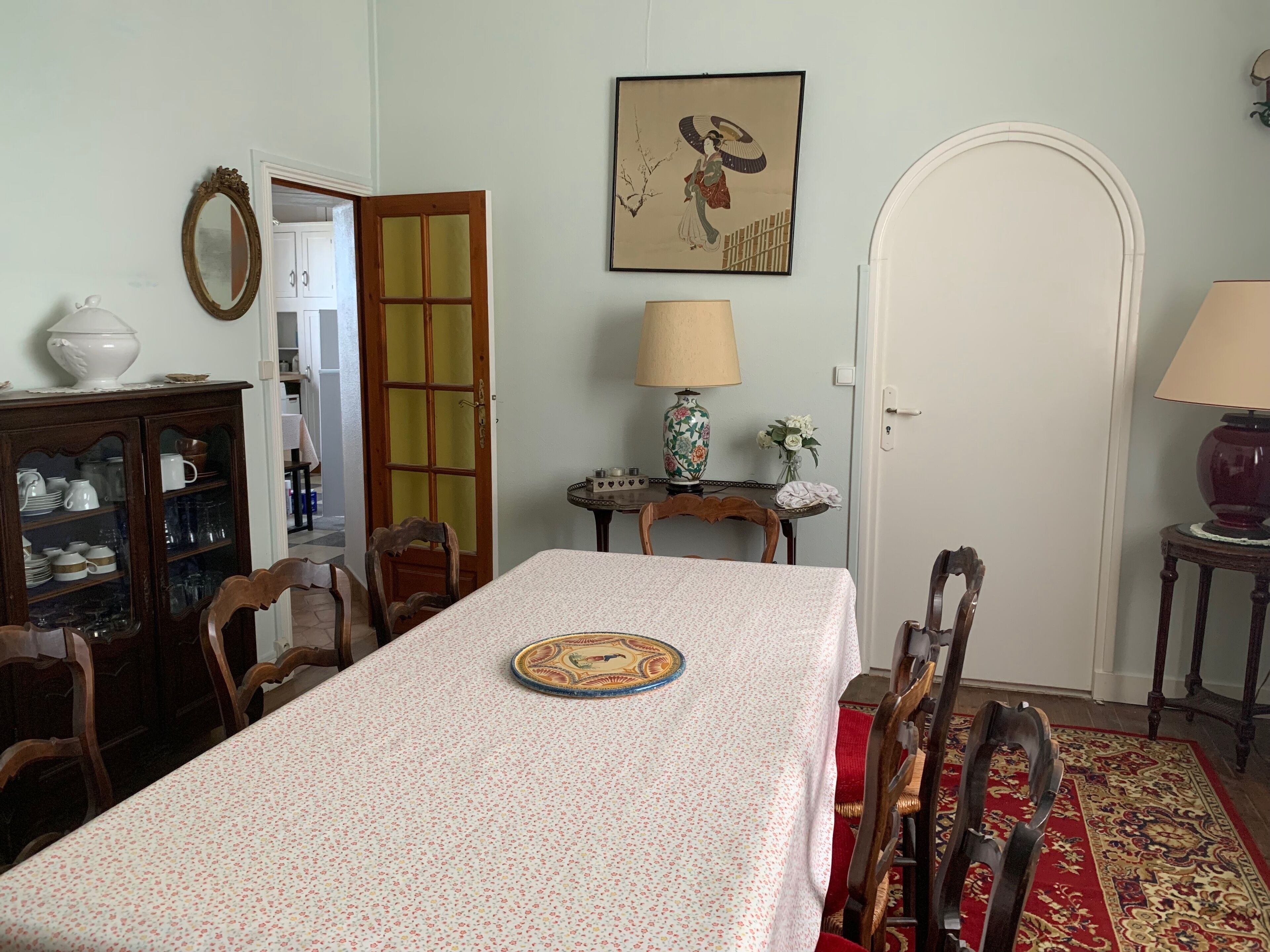 A dining room with a table set for four, surrounded by wooden chairs. A cabinet with glass doors is to the left, and a door with a glass panel is visible in the background.
