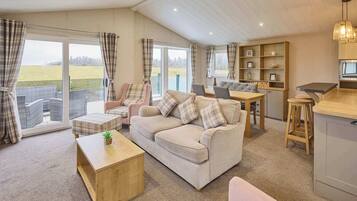 A cozy living room with a sofa, armchairs, a coffee table, and a dining area with a table and chairs. There is a bookshelf and a kitchen area with a counter and stools.
