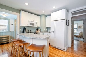 Une cuisine avec des meubles blancs, un plan de travail en granit, un four à micro-ondes encastré, un réfrigérateur et des tabourets en bois.