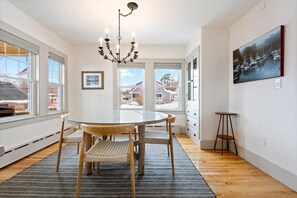 A dining area with a round glass table, wooden chairs, a chandelier, and a view of houses through large windows.