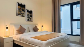Un lit bien fait, avec une literie blanche et marron, deux cadres photos accrochés au mur, une table de chevet avec une lampe, et une grande fenêtre avec des rideaux.