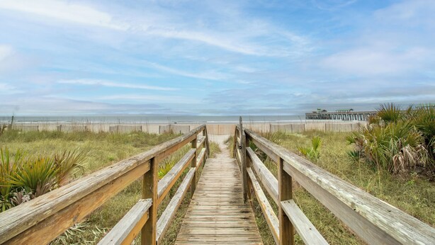 A wooden boardwalk leading to a beach with a wooden pier in the distance. - Oceanfront 30+ day rental! (Folly Beach)