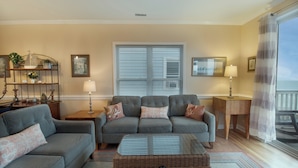 A living room with a grey sofa set, a wicker chest, and a window with blinds. - Oceanfront 30+ day rental! (Folly Beach)
