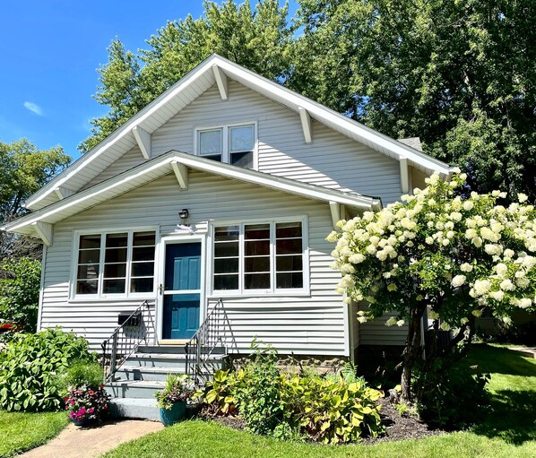 A single-story house with a front porch, a blue door, and a small front yard with various plants and a tree with white flowers. - Pleasant Stay in Pella (Pella)