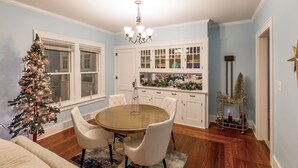 A dining area with a round wooden table, white upholstered chairs, a chandelier, and a Christmas tree. - Pleasant Stay in Pella (Pella)