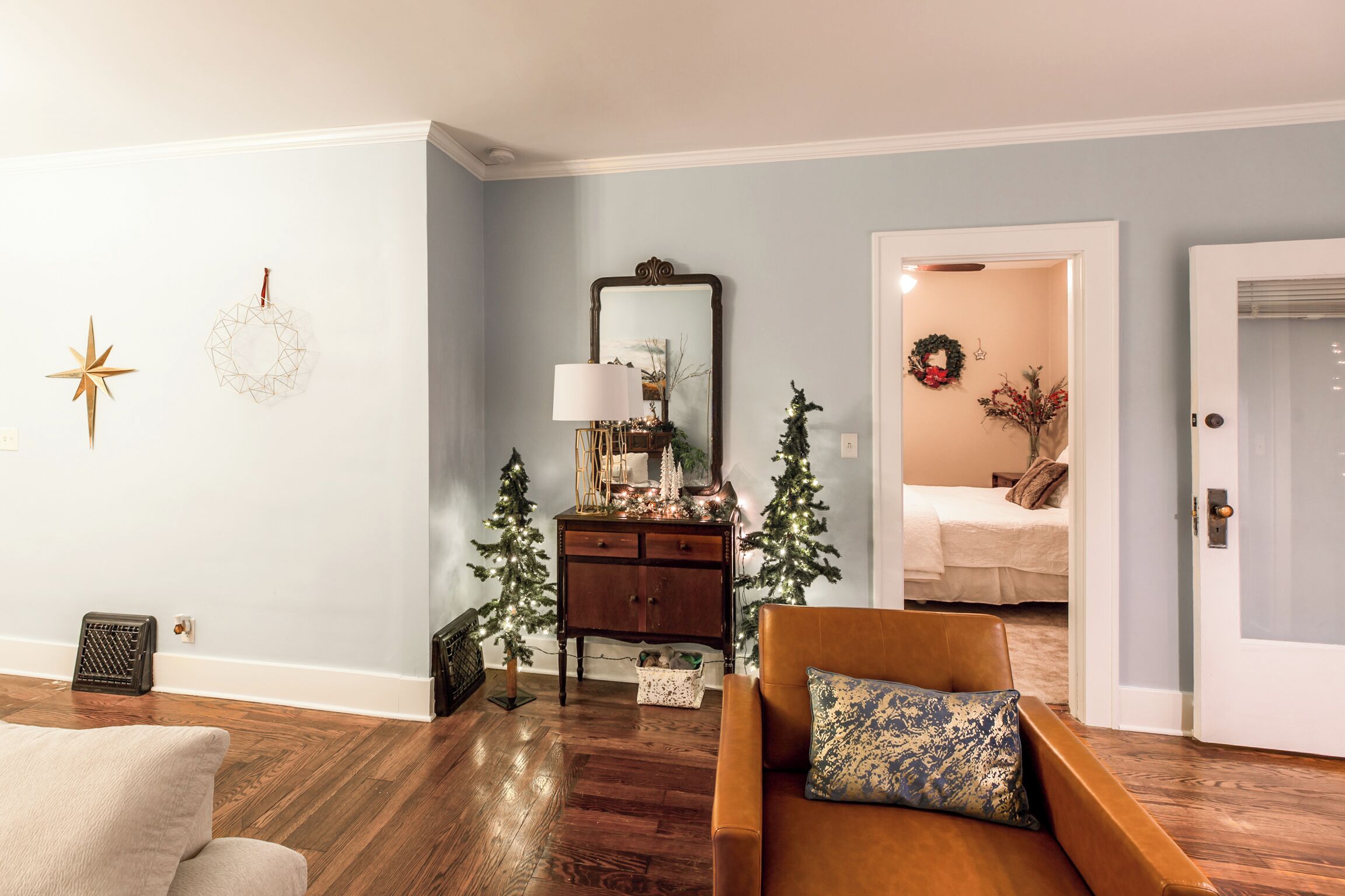 Una sala de estar con piso de madera, un sofá marrón, una mesita con una lámpara y dos árboles de Navidad.