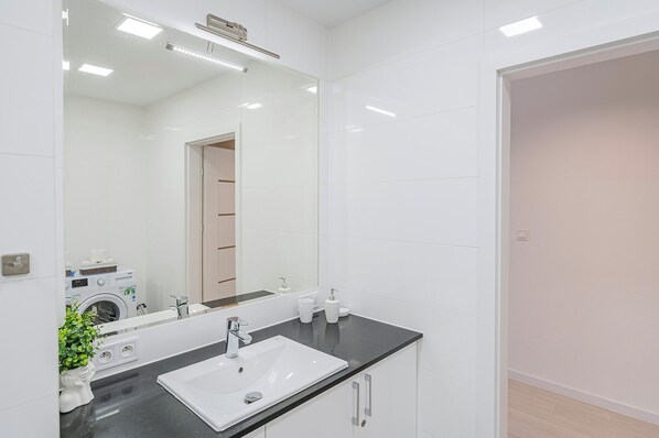 A modern bathroom with a large mirror, white tiles, a black countertop, a sink, and a washing machine visible in the reflection.