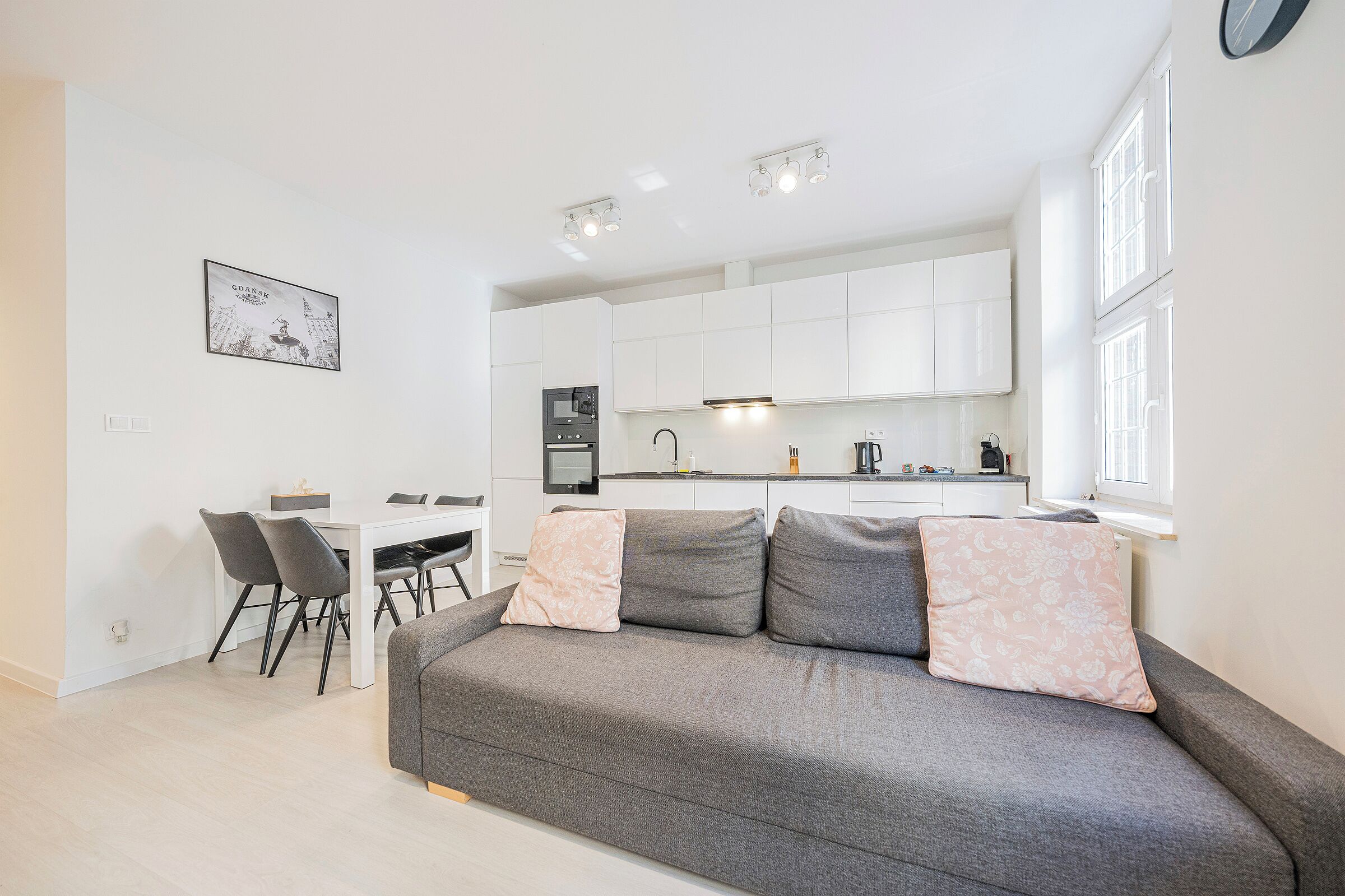 A modern apartment with a grey sofa, a dining table with chairs, a kitchen area, and a framed picture on the wall.