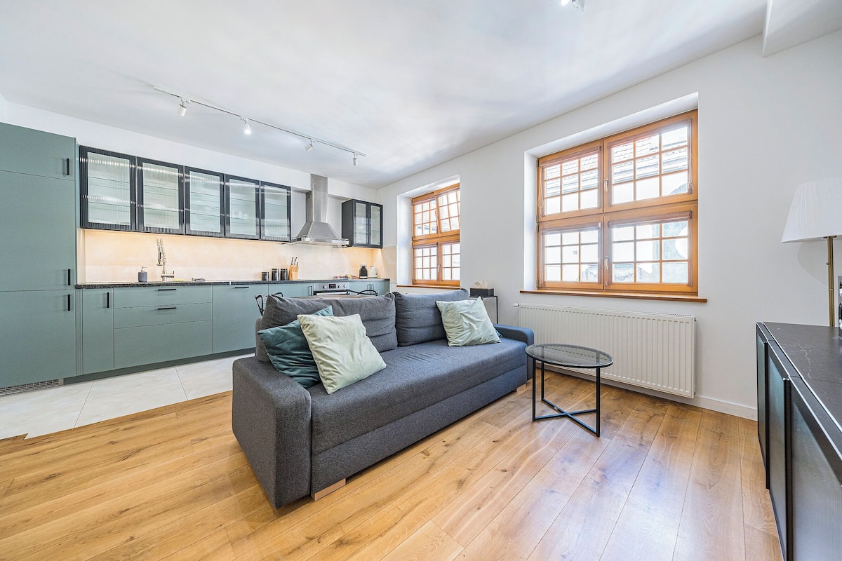 A modern kitchen with grey cabinets, a grey sofa, and a small table. There are large windows with wooden frames and a radiator below them.