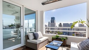 Un balcon moderne offrant une vue sur la ville, doté d’une porte vitrée, d’un coin salon confortable et d’une petite table.