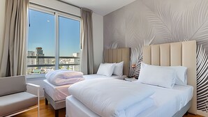 Une chambre d’hôtel moderne dotée d’un grand lit, d’un lit plus petit, d’un canapé et offrant une vue sur la ville grâce à de grandes fenêtres.