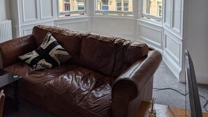A corner room with a brown leather sofa, white paneled walls, and large windows offering a view of buildings outside. - Traditional Victorian Edinburgh Flat Central Location, Bright and Airy (Edinburgh)