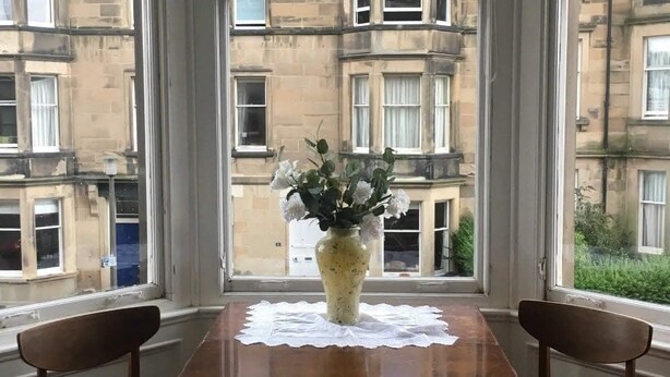 A dining area with a wooden table and two chairs, a vase with flowers, and large windows with a view of a building. - Traditional Victorian Edinburgh Flat Central Location, Bright and Airy (Edinburgh)