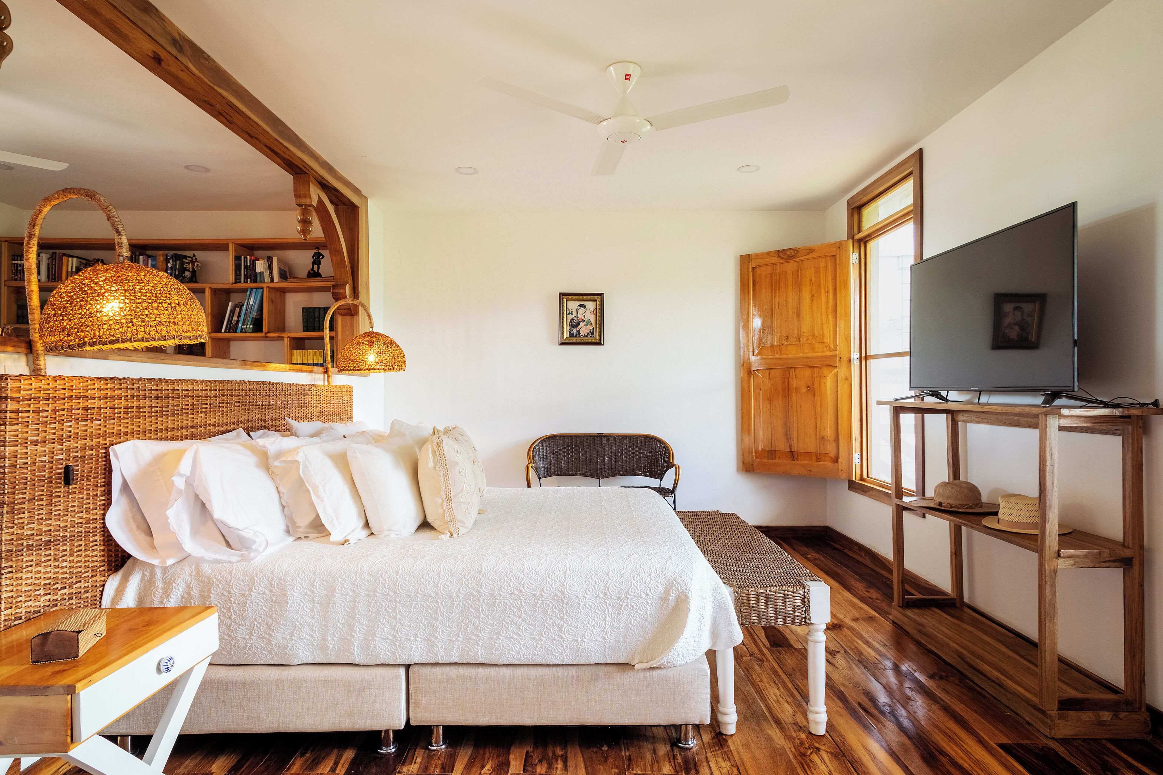 Ein Schlafzimmer mit einem großen Bett, einem Holz-Kopfteil, einem Deckenventilator, einem Fernseher auf einem Holzschrank und einem Bücherregal mit Büchern und Dekoration.