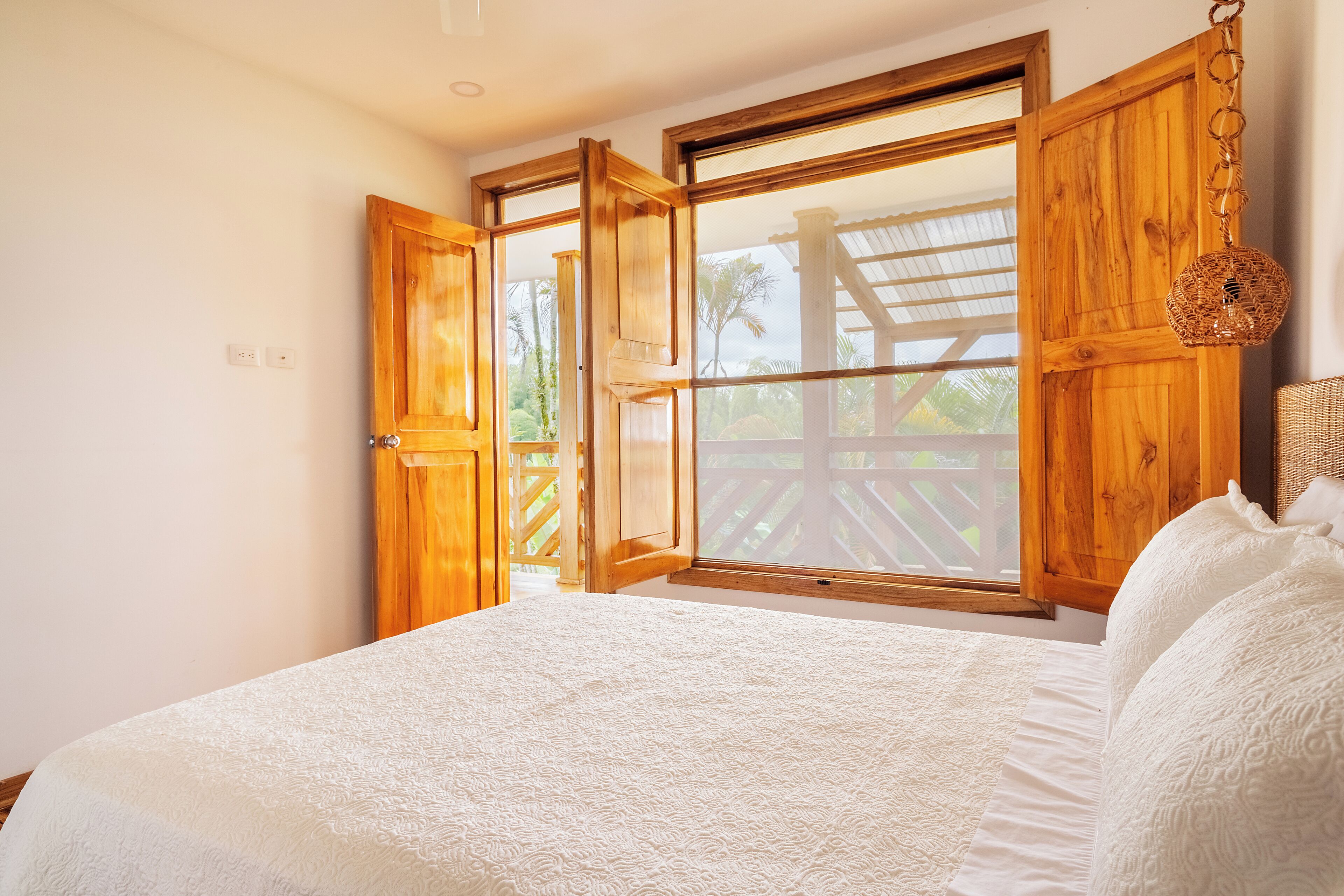 Une chambre avec un grand lit, une armoire en bois et une fenêtre donnant sur une terrasse en bois et des palmiers.