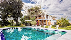 A swimming pool with a tiled deck, surrounded by white lounge chairs and a two-story building with a balcony. - Casa de Campo el Edén (Armenia)