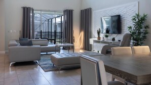 A modern living room with a sofa, ottoman, dining table, and chairs. There is a flat-screen TV mounted on the wall and a potted plant on the right. - 5BR house with game room near Disney (Kissimmee)
