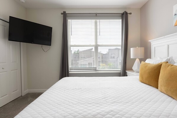 A bedroom with a large bed, a flat-screen TV mounted on the wall, a window with blinds, a bedside lamp, and a view of buildings outside.