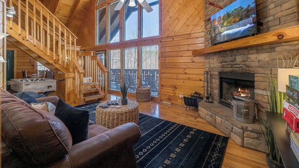 A cozy cabin interior with a fireplace, wooden walls, a ceiling fan, and a large window overlooking a deck.