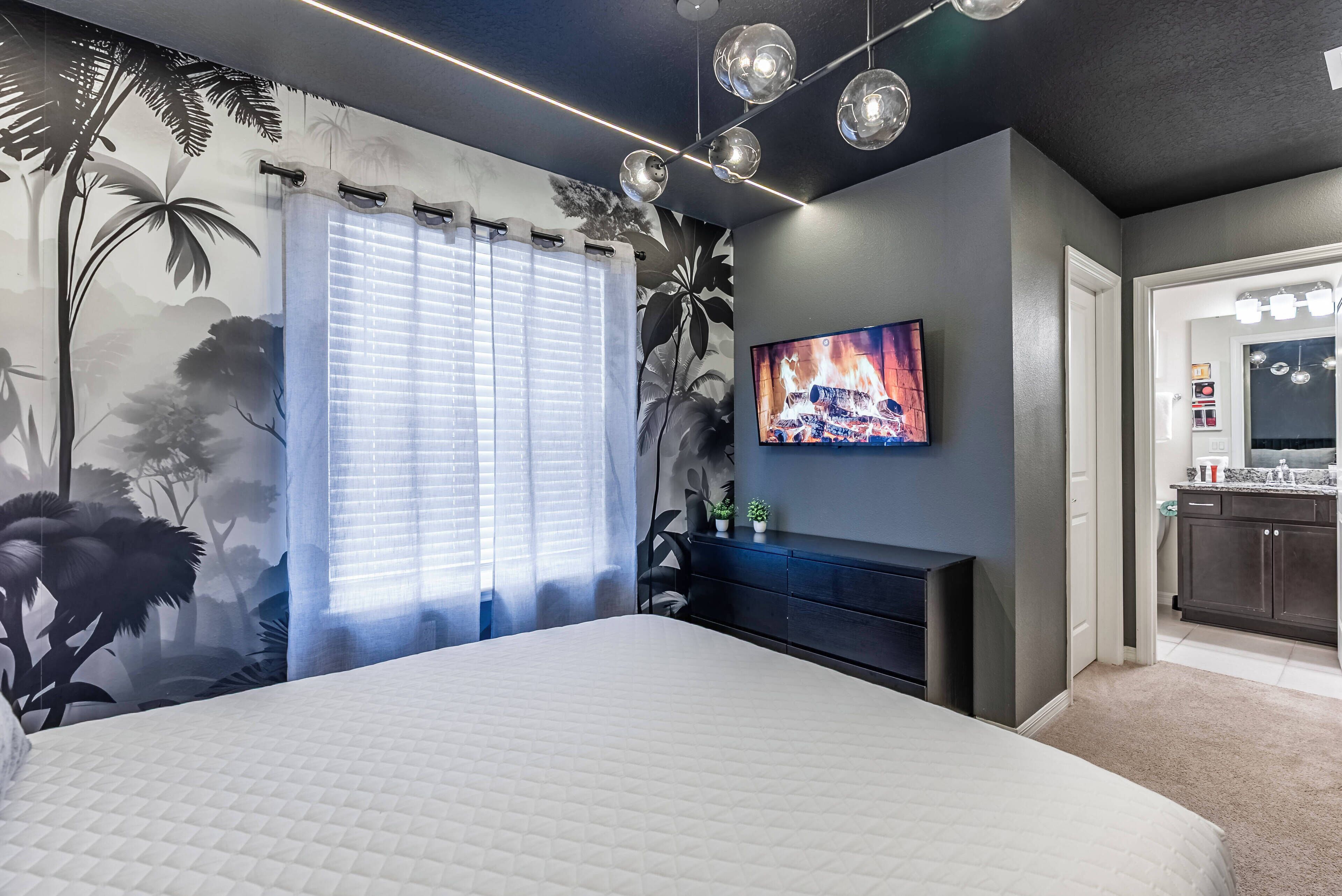 Ein Schlafzimmer mit einem großen Bett, einem an der Wand montierten Fernseher, einer dunklen Kommode und einem Fenster mit Jalousien.
