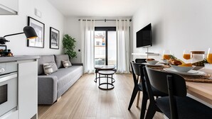 Una sala de estar moderna con un sofá gris, una mesa de comedor con platos y vasos, un televisor montado en la pared y una ventana con cortinas blancas.