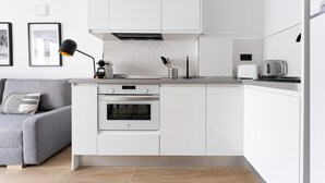 Una cocina moderna con armarios blancos, horno empotrado, cafetera y un sofá gris en la sala de estar contigua.