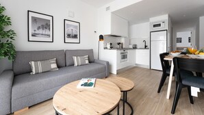 Una sala de estar moderna con un sofá gris, dos mesitas de madera y un comedor con una mesa blanco y negro. Hay cuadros en la pared y una planta a la izquierda.