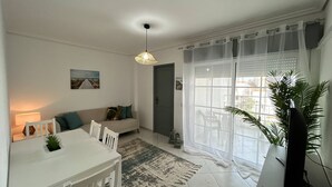 Een woonkamer met een bank, een eettafel, een lamp en een groot raam met dunne gordijnen.