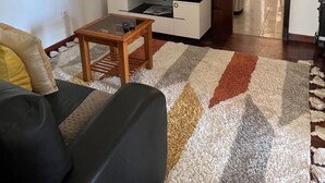 Olohuoneessa on musta sohva, puinen sohvapöytä, valkoisessa kaapissa oleva taulutelevisio ja kuviollinen matto.