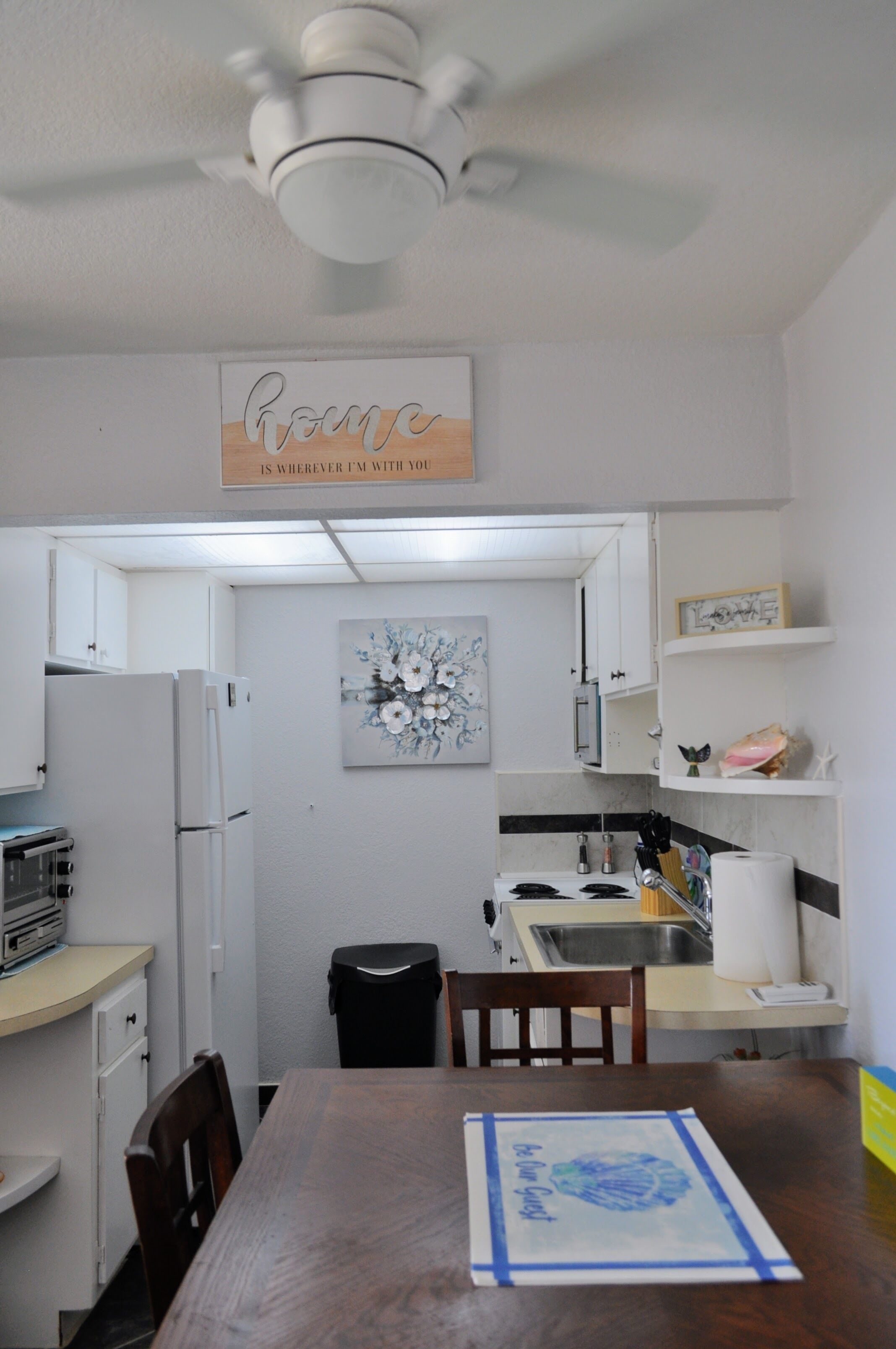 Una cucina compatta con mobili bianchi, un frigorifero, un lavandino e un tavolo con sedie. Si nota una ventola a soffitto, un'opera d'arte appesa al muro e un cartello sopra la porta.