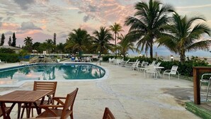 A pool area with lounge chairs and tables, surrounded by palm trees and a clear sky. - Coral Beach Retreat. Garden view. Steps from pool & beach. 1 mile to Port Lucaya (Freeport)