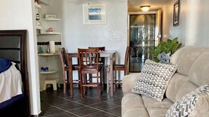 A living room with a beige sofa, patterned pillows, a dining area with wooden chairs, a small table, and a kitchen visible through a glass door. - Coral Beach Retreat. Garden view. Steps from pool & beach. 1 mile to Port Lucaya (Freeport)