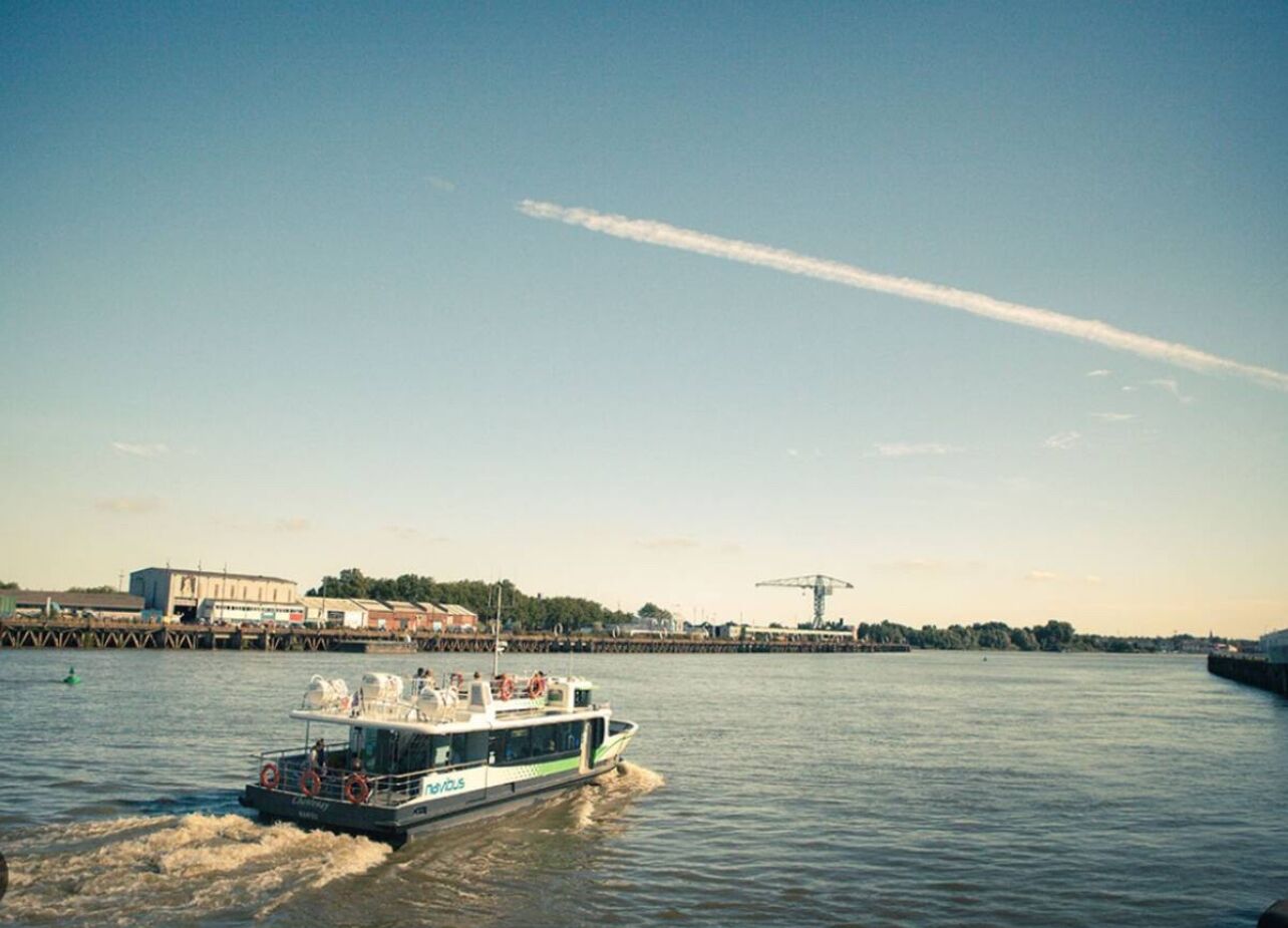 A riverboat with passengers, docked near a pier with industrial buildings and a crane in the background.