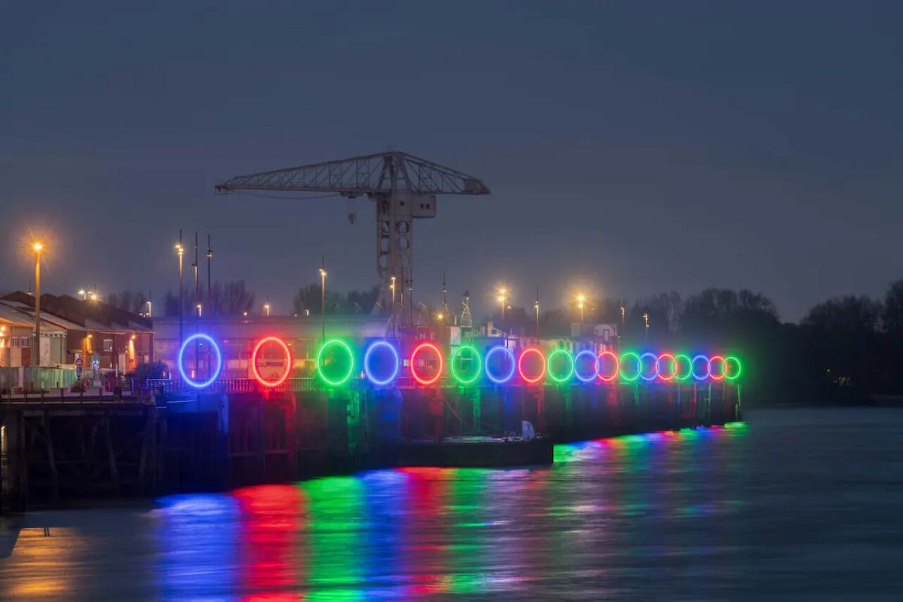 Ein Uferbereich mit Anlegesteg und Kran, beleuchtet durch farbige Lichter, die sich im Wasser spiegeln.