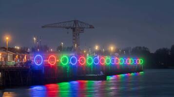 Ein Uferbereich mit Anlegesteg und Kran, beleuchtet durch farbige Lichter, die sich im Wasser spiegeln.