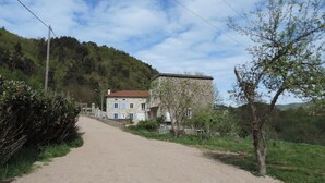 Un camino rural que lleva a una casa de piedra con techo rojo, rodeada de vegetación y colinas.