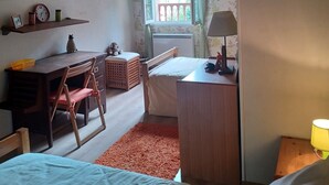 A cozy room with a wooden ceiling, a bed with a green pillow, a desk with a chair, a small table with a lamp, and a window with a view of greenery.