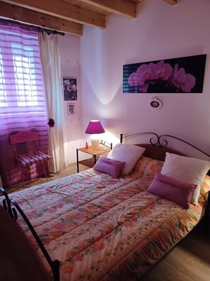 Ein Schlafzimmer mit Blumenmuster-Bettdecke, einem Nachttisch mit Lampe, einem Sessel und einem Fenster mit Vorhängen.