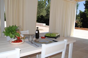 An outdoor dining table with a bowl of tomatoes, a bottle, and a green object, possibly a food processor, under a covered area with curtains.