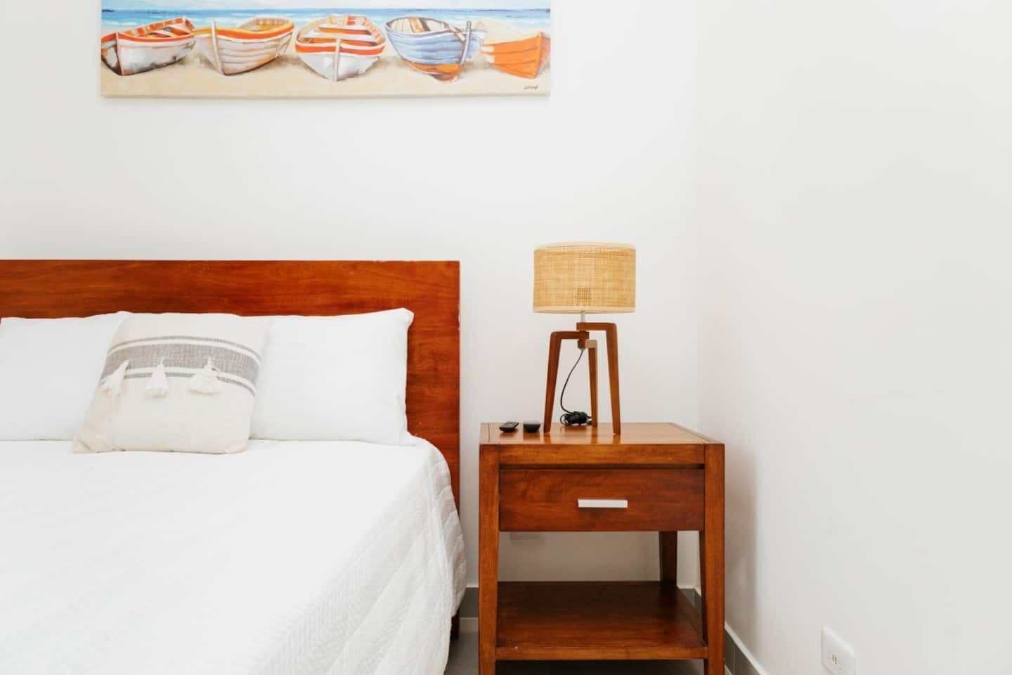 A bed with white bedding, a wooden nightstand with a lamp, and a painting of boats on the wall.