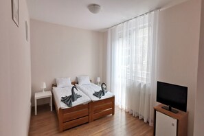 A bedroom with two beds, a bedside table, a TV stand, and a window with curtains.