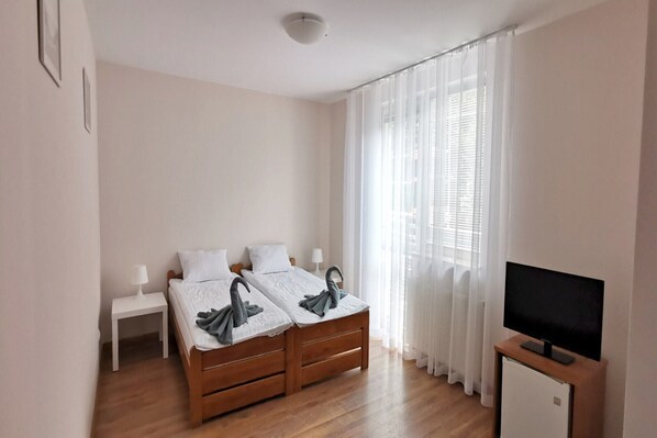 A bedroom with two beds, a bedside table, a TV stand, and a window with curtains.