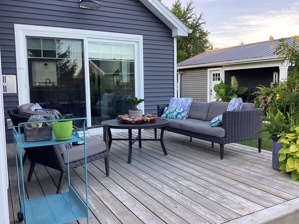 A deck with a sofa, table, and chairs, surrounded by potted plants and a house with a dark siding exterior.