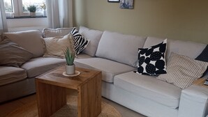 A living room with a beige sofa, a wooden coffee table, and a woven rug. There are two side tables, one with a potted plant. The room has a large window with curtains, and the walls are decorated with framed pictures. - TKS HOME Am Weiherplatz In the heart of the Westerwald (Norken)