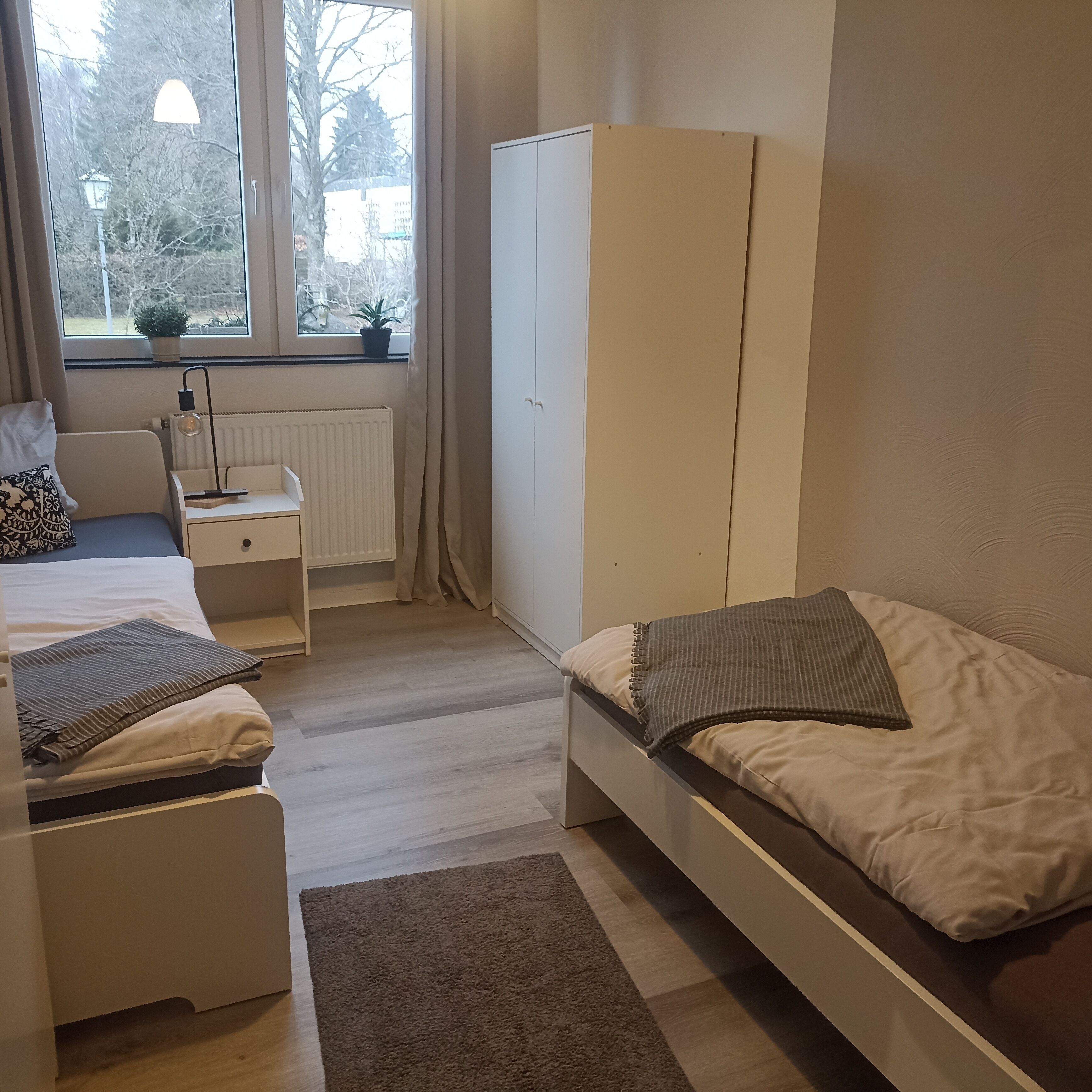 Quarto com cama, escrivaninha, guarda-roupa e janela com vista para árvores.