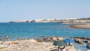 A rocky beach with people sunbathing and swimming, a clear blue sea, and a distant cityscape.
